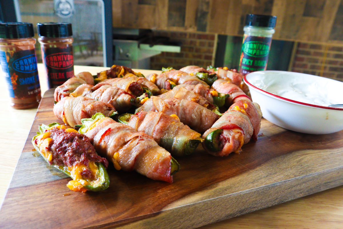 Bacon-wrapped stuffed jalapeños arranged on a wooden cutting board, with a small white dipping bowl and carnivore companion meat seasoning in the background