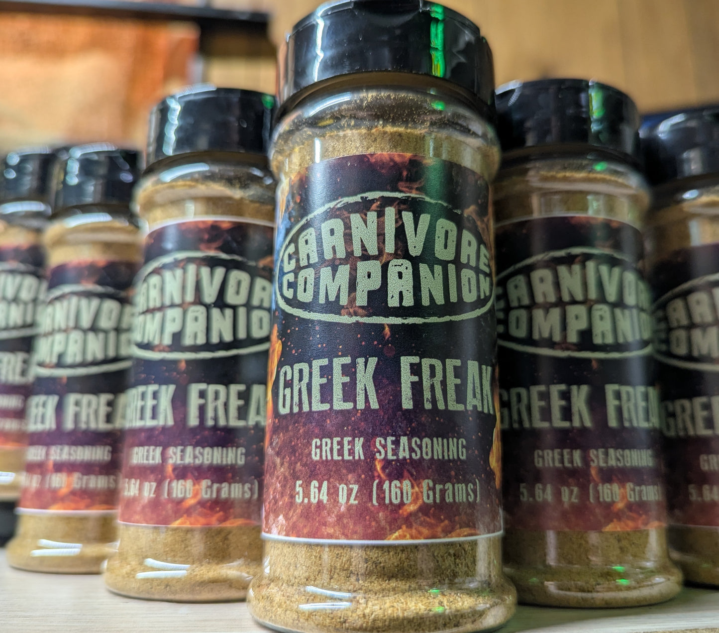 A close-up of several spice jars labeled 'Carnivore Companion Greek Freak Greek Seasoning' standing on a shelf. The center jar is in sharp focus, with other jars blurred in the background.