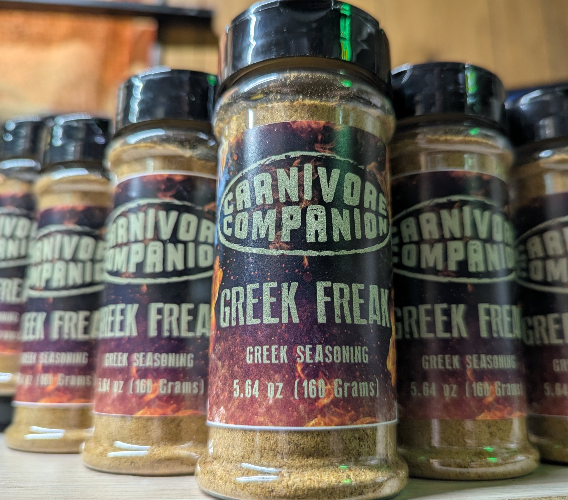 A close-up of several spice jars labeled 'Carnivore Companion Greek Freak Greek Seasoning' standing on a shelf. The center jar is in sharp focus, with other jars blurred in the background.